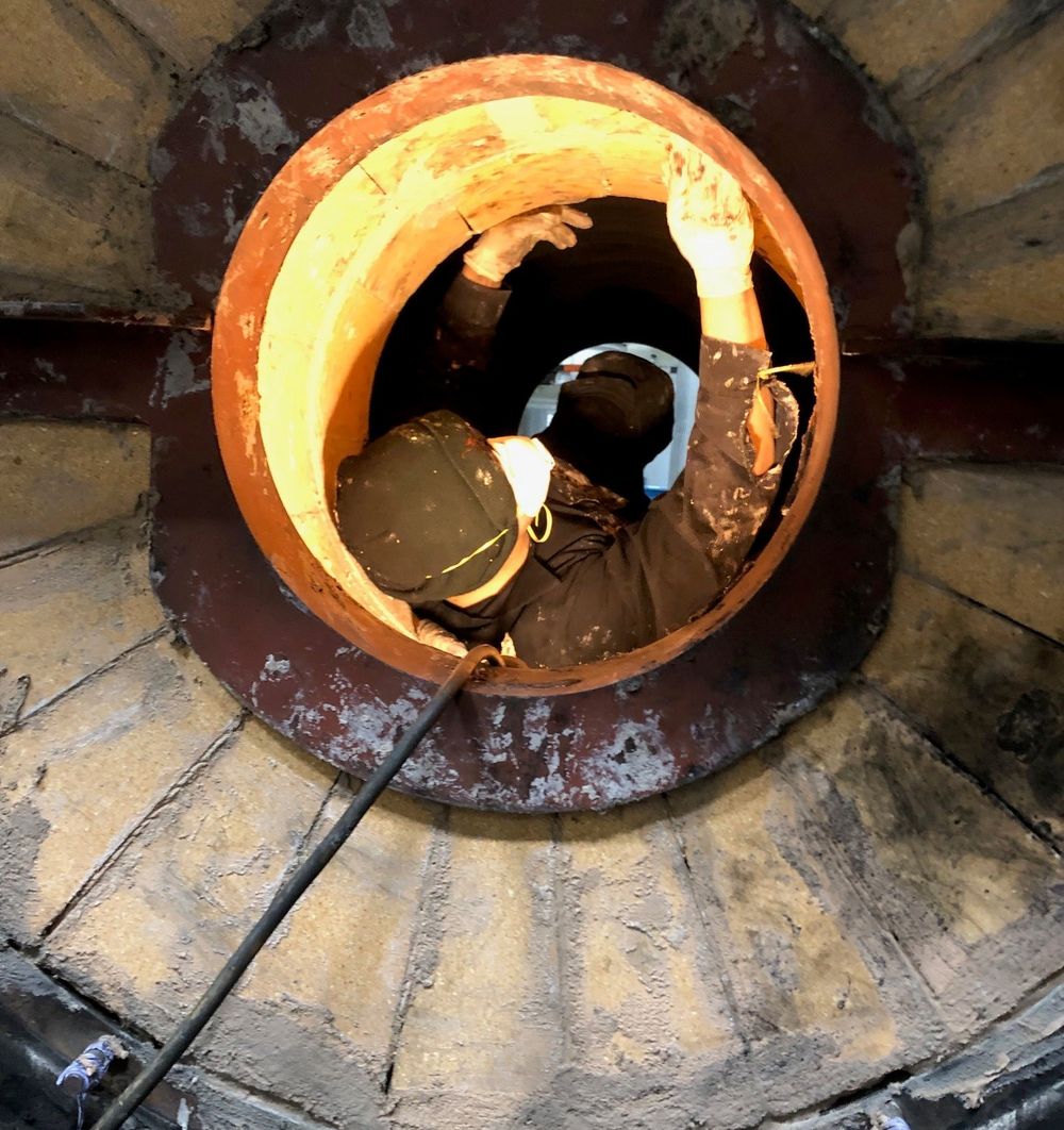 Technician cleaning the fireside of a commercial boiler furnace tube, viewed through the refractory-lined rear chamber